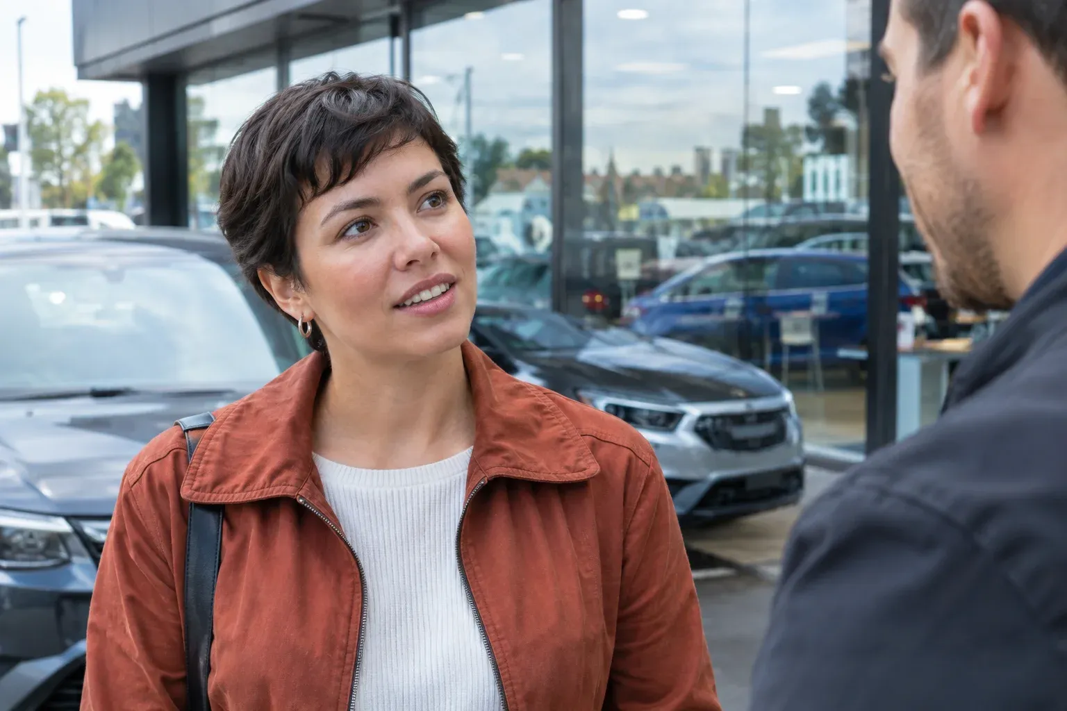A woman with short dark hair wearing a rust jacket in conversation outside a dealership, glass reflects cars
