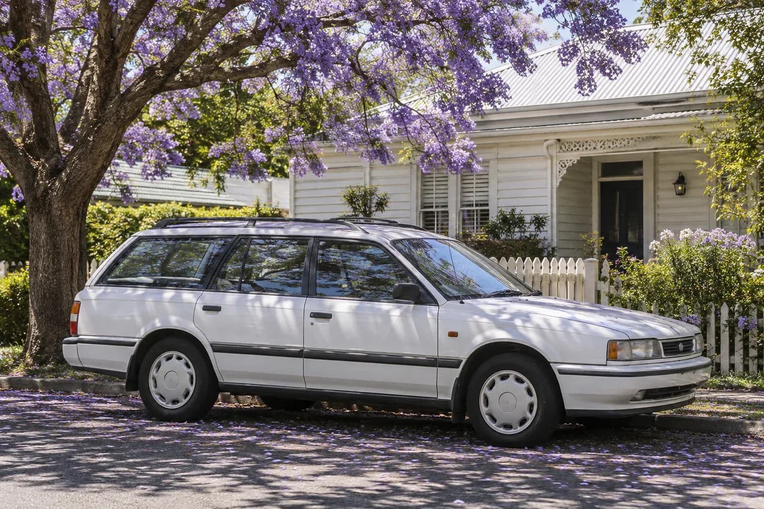 A well-kept older white station wagon parked beside a weatherboard home, afternoon shadows from a jacaranda