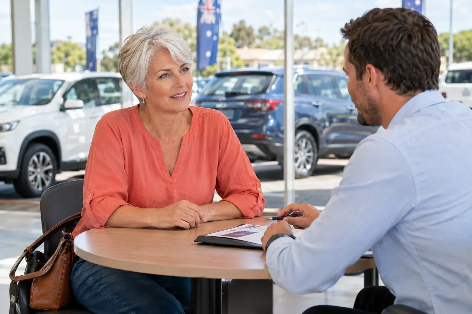 A woman with short silver hair in a coral blouse and a male adviser listening across a small round table, car forecourt visible beyond