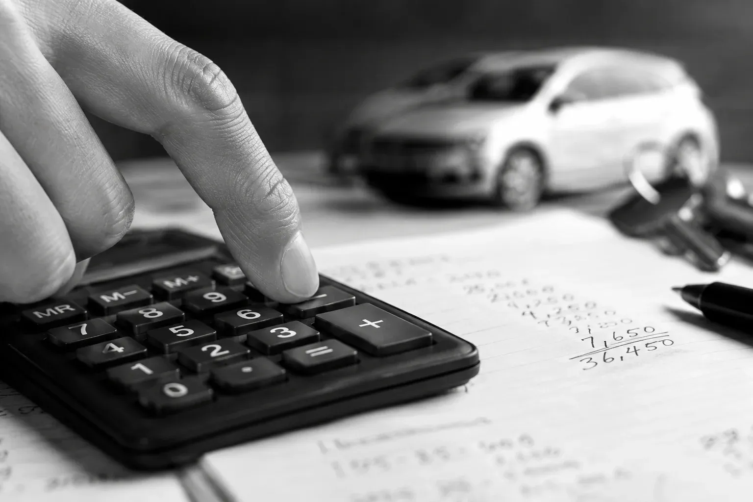 Fingers pressing a black calculator beside handwritten arithmetic blurred beyond focus
